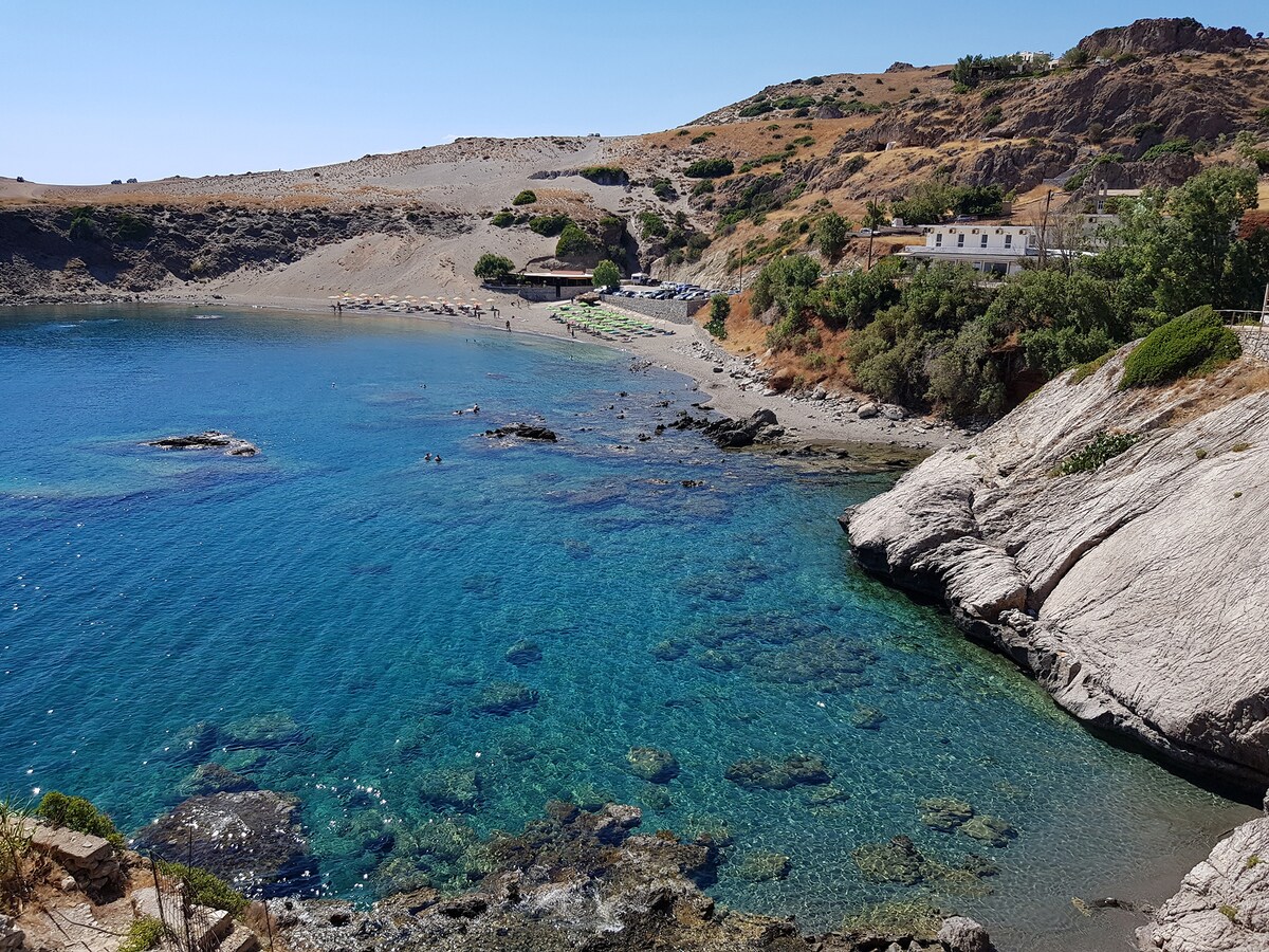 The clear blue water of the cove is surrounded by rocky shores and sandy areas. A gently sloping hillside with sparse vegetation is visible in the background. Several people can be seen enjoying the beach, while the coastline curves gracefully into the distance.