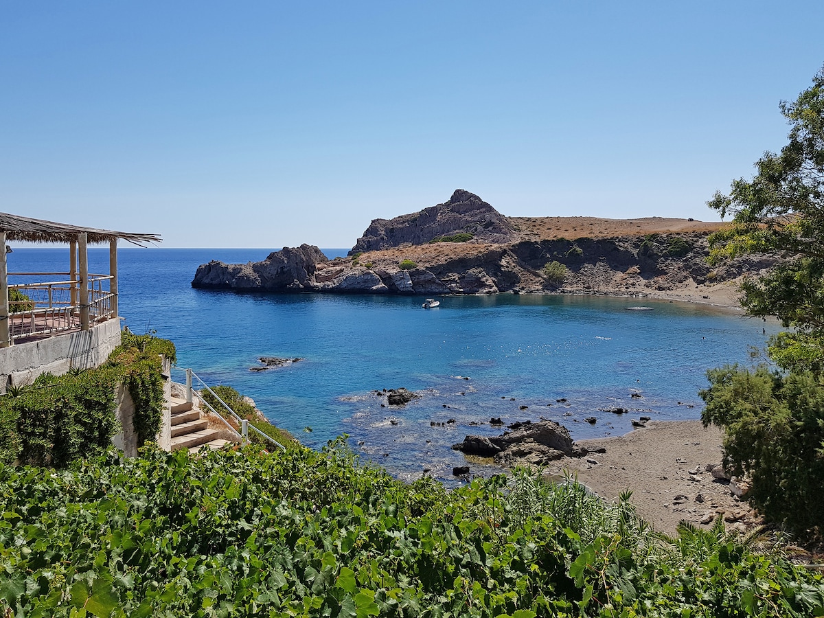 A serene cove is depicted with clear blue waters gently lapping at the rocky shore. A small boat can be seen floating in the distance. The surrounding landscape features dry hills and patches of greenery, under a bright, cloudless sky.