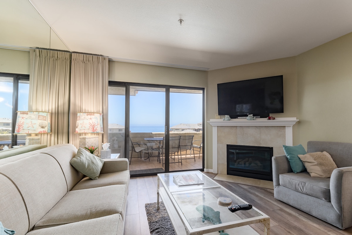 The living room features a comfortable couch and a recliner, with large windows providing ocean views. A flat-screen TV is mounted above a gas fireplace, while a coffee table sits on a soft area rug. Sliding glass doors open to a private balcony.
