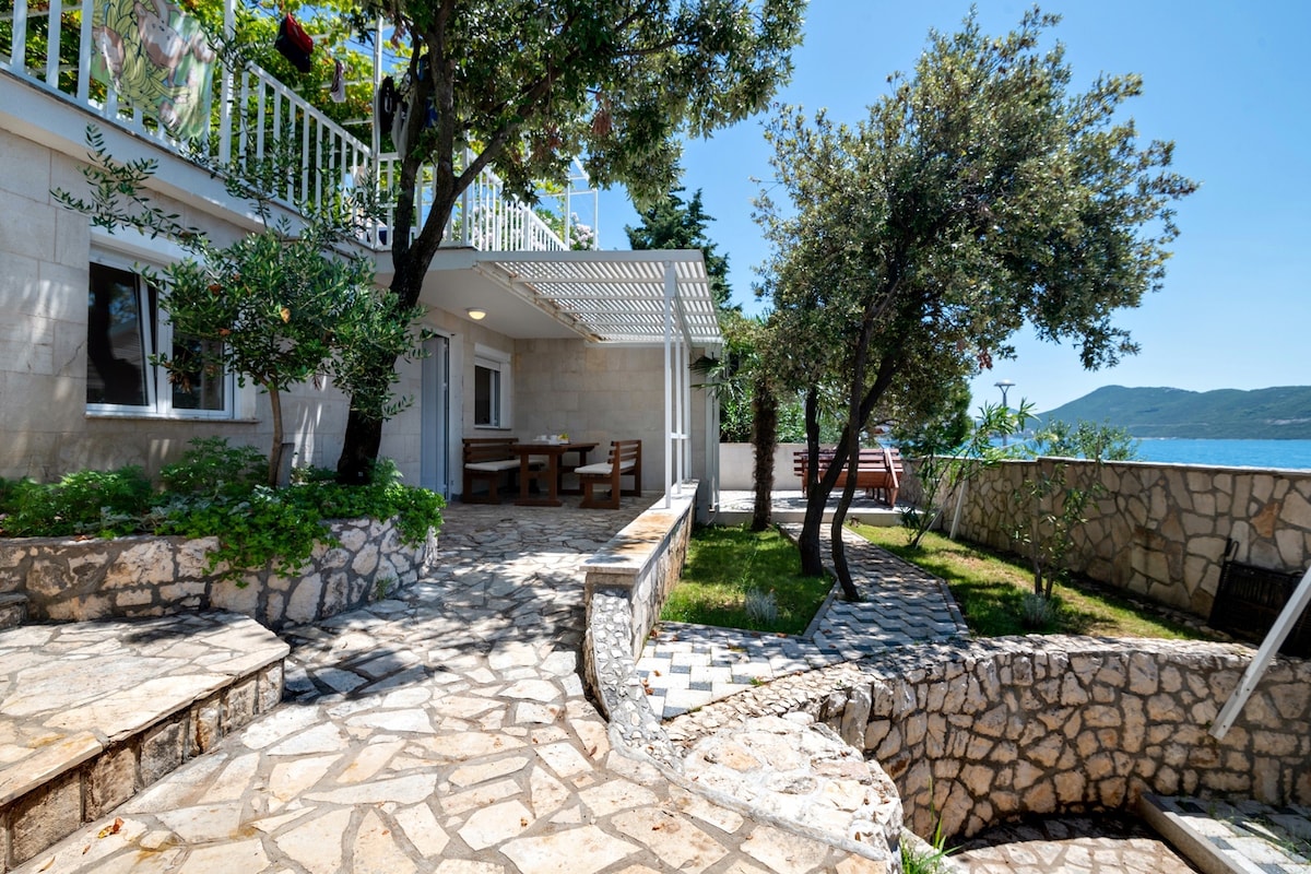 A stone pathway leads to a terrace surrounded by lush greenery. The outdoor area features wooden dining furniture and views of the seaside. Sunlight casts gentle shadows on the stone surfaces, highlighting the natural texture of the landscape.