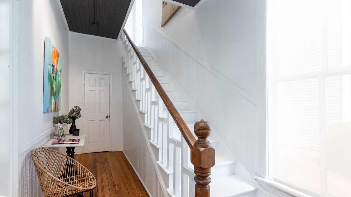 A well-lit hallway features white walls and wooden floors, leading to a staircase with a natural wood railing. A modern artwork adorns the wall, while a decorative chair provides a touch of charm beside a small table.