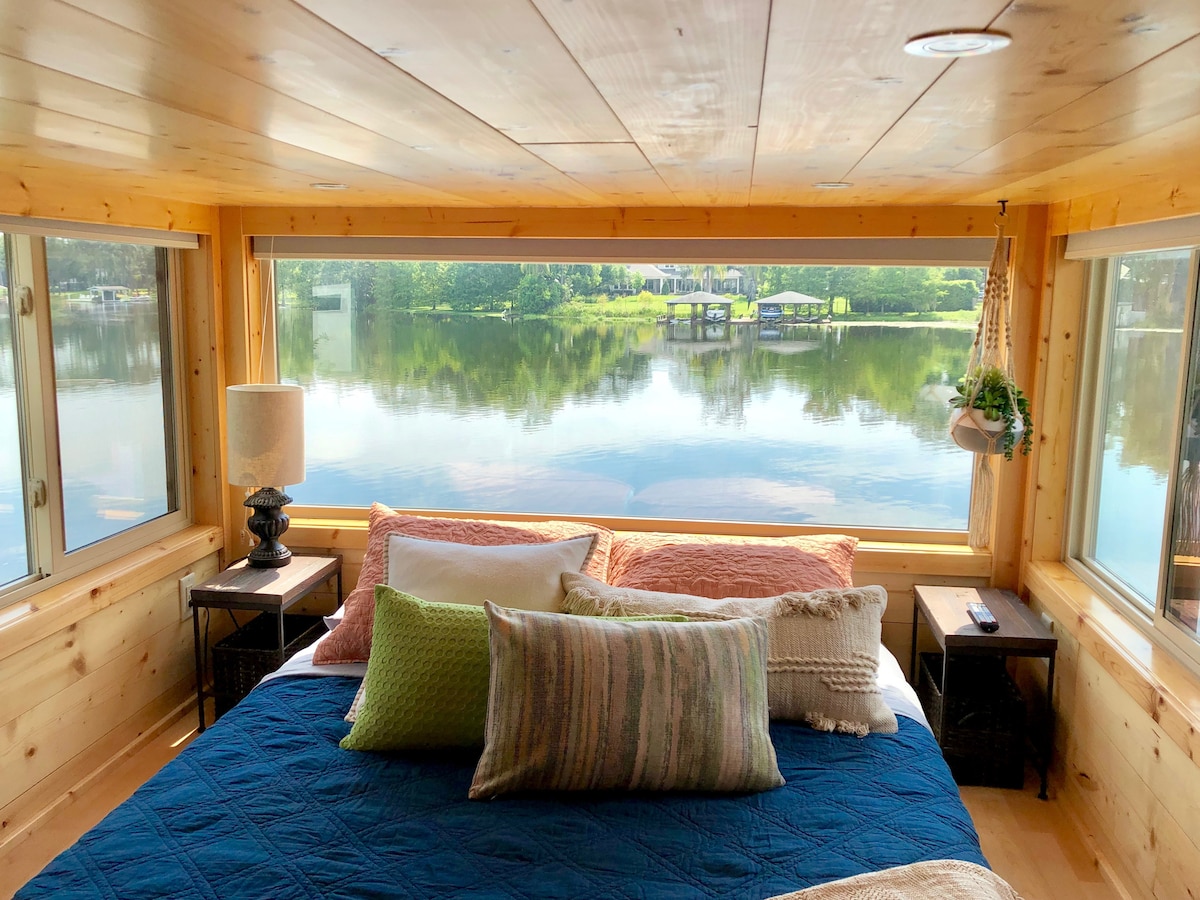 A cozy sleeping area features a spacious bed with a dark blue quilt and an array of colorful pillows. Large picture windows frame a serene view of the lake and surrounding greenery, while two lamps provide soft lighting on either side of the bed.