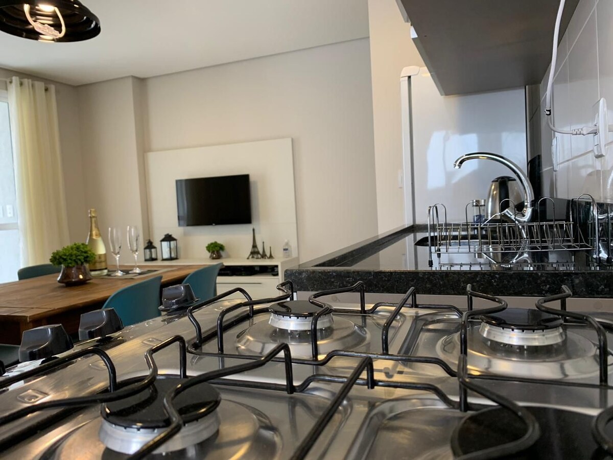 A modern kitchen area is visible, featuring a gas cooktop with multiple burners. The sleek black countertop provides a contrasting backdrop for stainless steel appliances. In the background, a dining area is suggested with a wooden table and chairs, along with a television mounted on the wall.