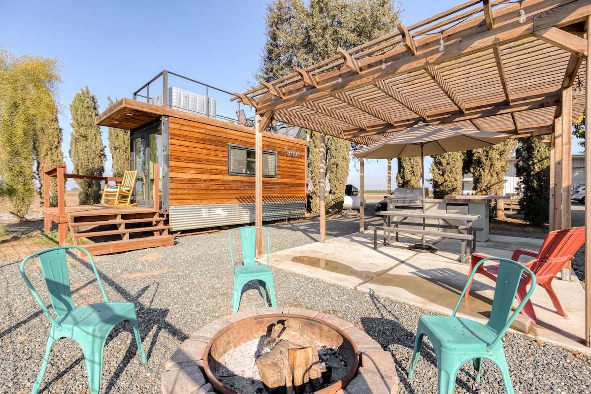 An outdoor space is shown featuring a fire pit surrounded by colorful chairs. A shaded dining area with a long table and umbrellas is visible, alongside a tiny home with wooden siding and a rooftop deck. Greenery frames the scene, enhancing the rural setting.