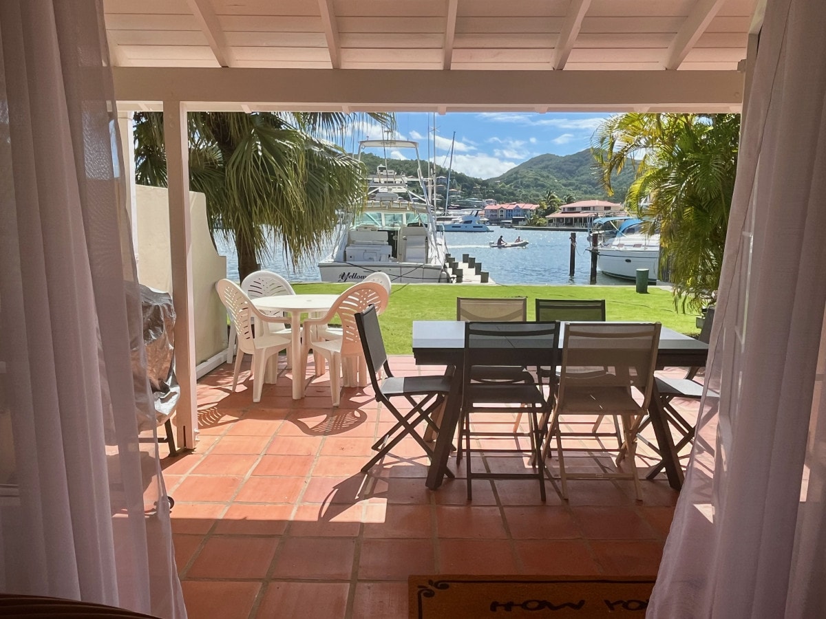 An outdoor patio area is visible, featuring a dining table and chairs facing waterfront views. The space is bordered by white curtains, allowing natural light to enter. Lush greenery and boats can be seen in the background, enhancing the tranquil atmosphere.