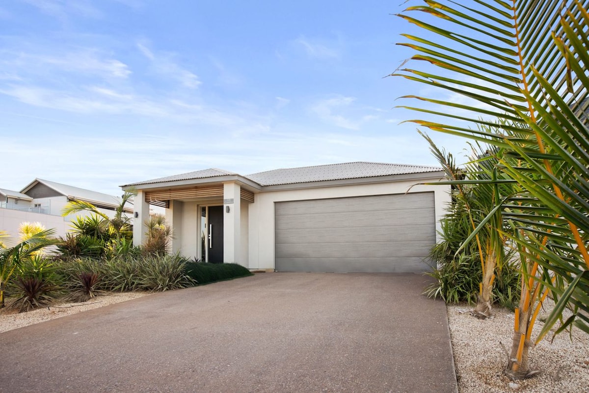 The exterior of the home features a modern façade with a spacious driveway lined by tropical plants. A double garage is visible, providing convenient parking in a serene setting. The property is surrounded by landscaped greenery, creating an inviting curb appeal.