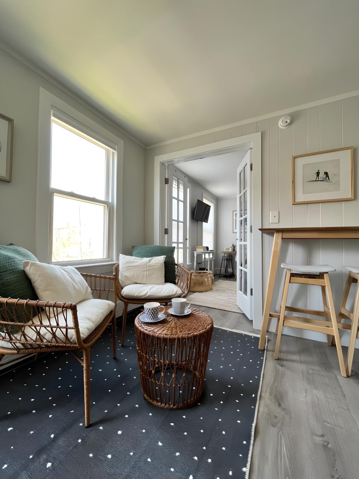 A cozy seating area is featuring two wicker armchairs with plush cushions, positioned near large windows that allow natural light to fill the space. A circular wooden side table holds decorative items, while a black and white patterned rug anchors the seating.