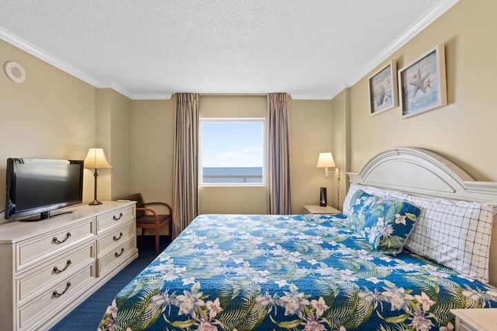 Svijetla i udobna spavaća soba s prekrivačem s cvjetnim uzorkom, prozorom s pogledom na more i šarmantnim uređenjem za opuštajući odmor.