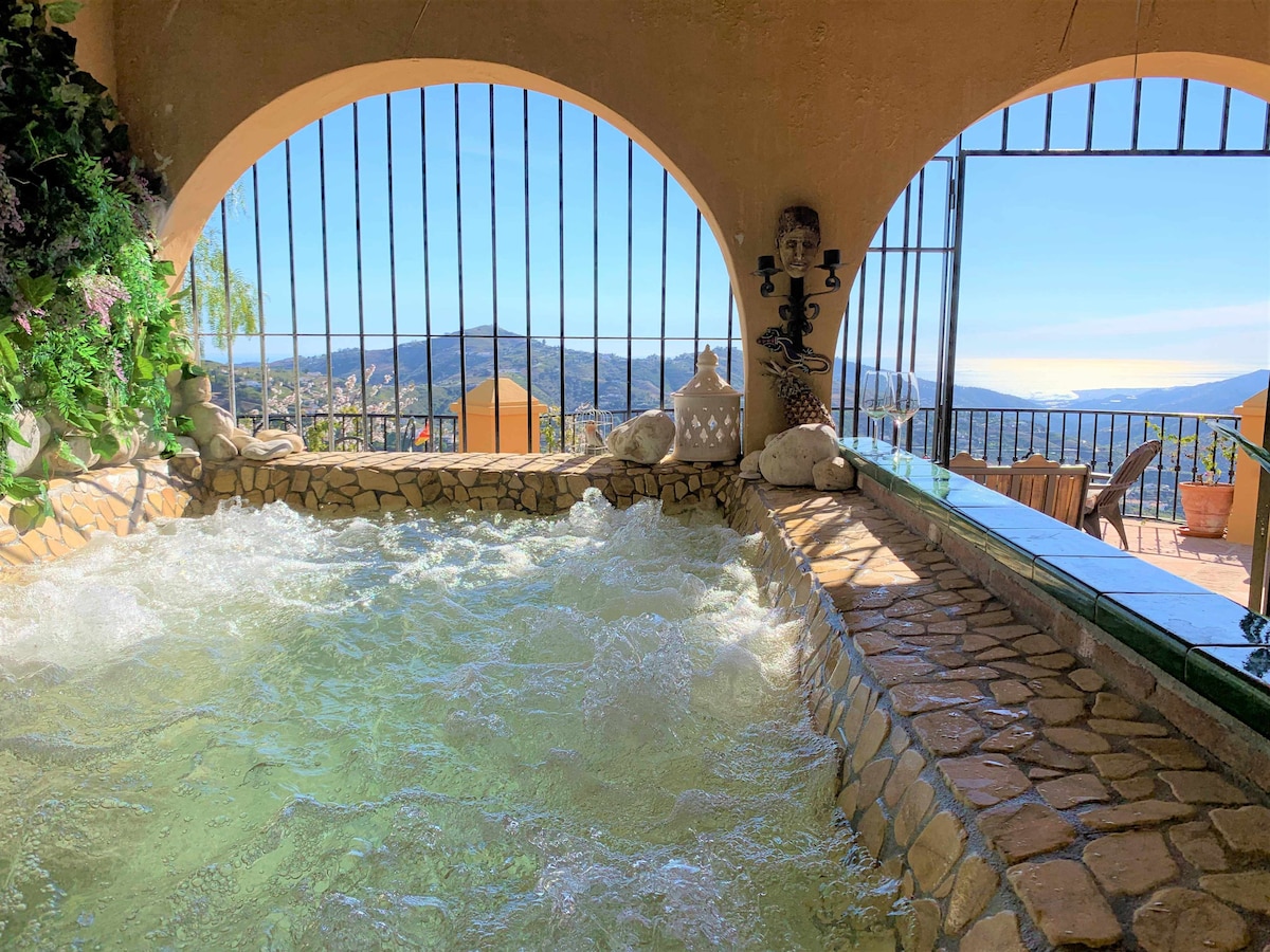 A serene jacuzzi is framed by stone archways, offering views of the surrounding mountains and coastline. The bubbling water is illuminated by natural light streaming through the bars. Lush greenery decorates the interior, creating a tranquil setting for relaxation.