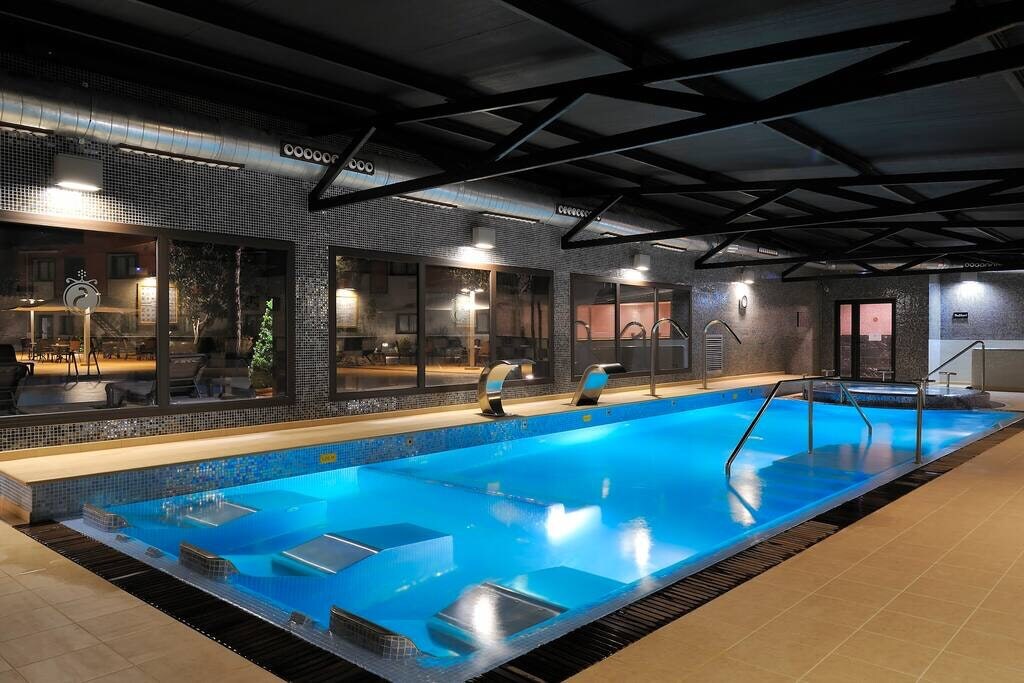An indoor pool area is showcased, featuring a well-maintained blue pool with built-in steps for easy access. Large windows allow natural light to illuminate the space, while sleek tile accents and metal beams provide a modern aesthetic.