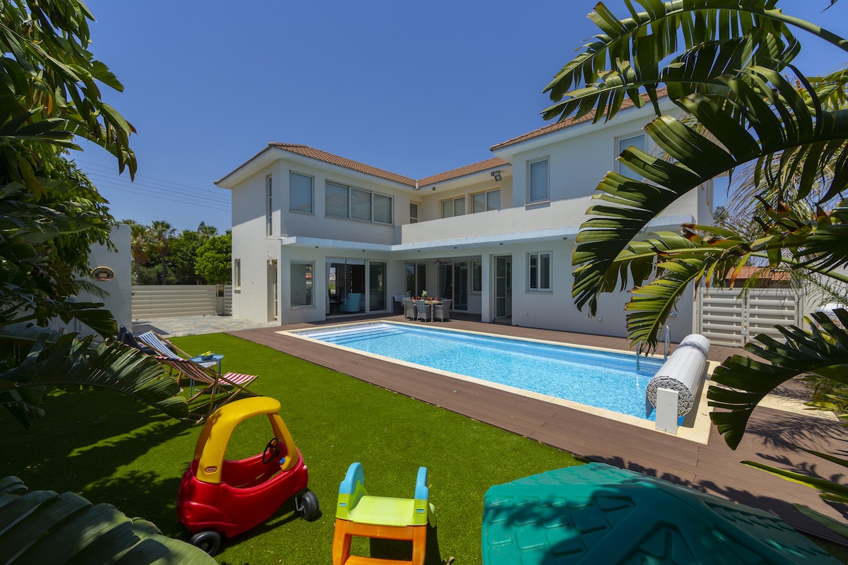 The exterior of a modern villa is shown, featuring a large private pool surrounded by a manicured lawn. Colorful children's toys are positioned on the grass, while the two-story structure with multiple windows reflects the bright blue sky.