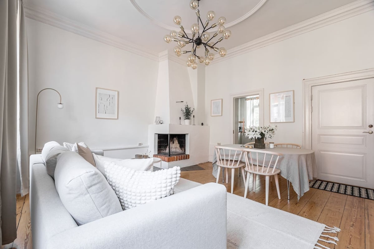 A spacious living room features a light grey couch adorned with textured cushions, a dining table with seating for four, and large windows that allow natural light to fill the space. A modern chandelier hangs above, with a neutral area rug and a fireplace adding warmth to the ambiance.