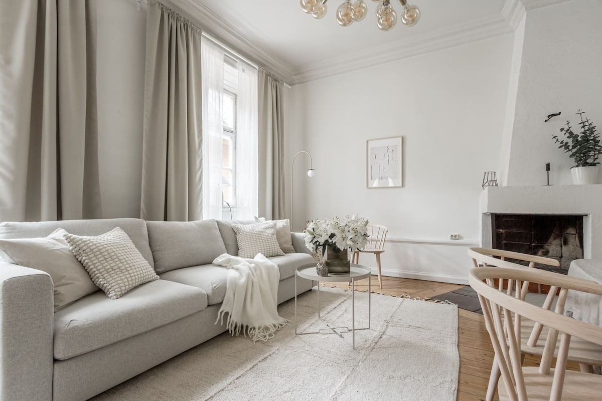 The living room features a light grey couch adorned with textured cushions and a decorative throw. Two wooden chairs and a small coffee table are arranged on a soft area rug. Natural light filters through large windows covered by sheer curtains.