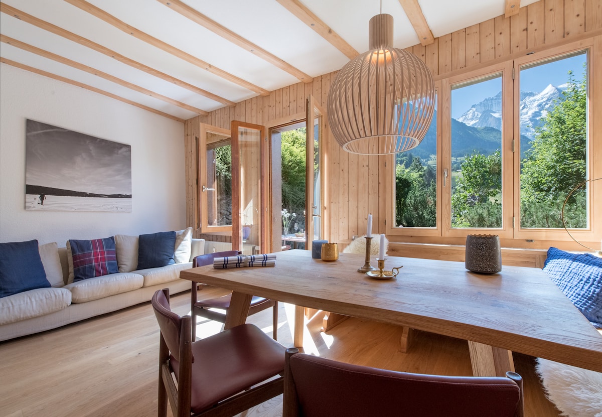 The open-plan living, dining, and kitchen area is characterized by wooden walls and beams, creating a warm atmosphere. Large windows provide natural light and scenic views of the surrounding mountains. A large wooden table is accompanied by brown chairs, while a comfortable sofa offers additional seating.
