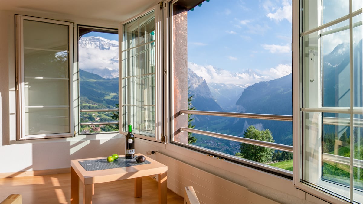 Large windows frame stunning panoramic views of the surrounding mountains and valleys. A small table with a bottle of wine and a lime is positioned in the light-filled corner, inviting guests to relax and enjoy the scenery.