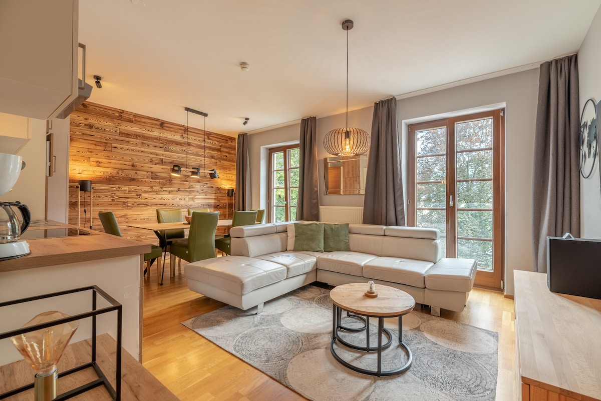 An open-plan living area features a modern sectional sofa with decorative cushions, a round coffee table, and a dining space with six chairs. Large windows provide natural light, and a wooden accent wall adds warmth. Light fixtures complement the contemporary design.