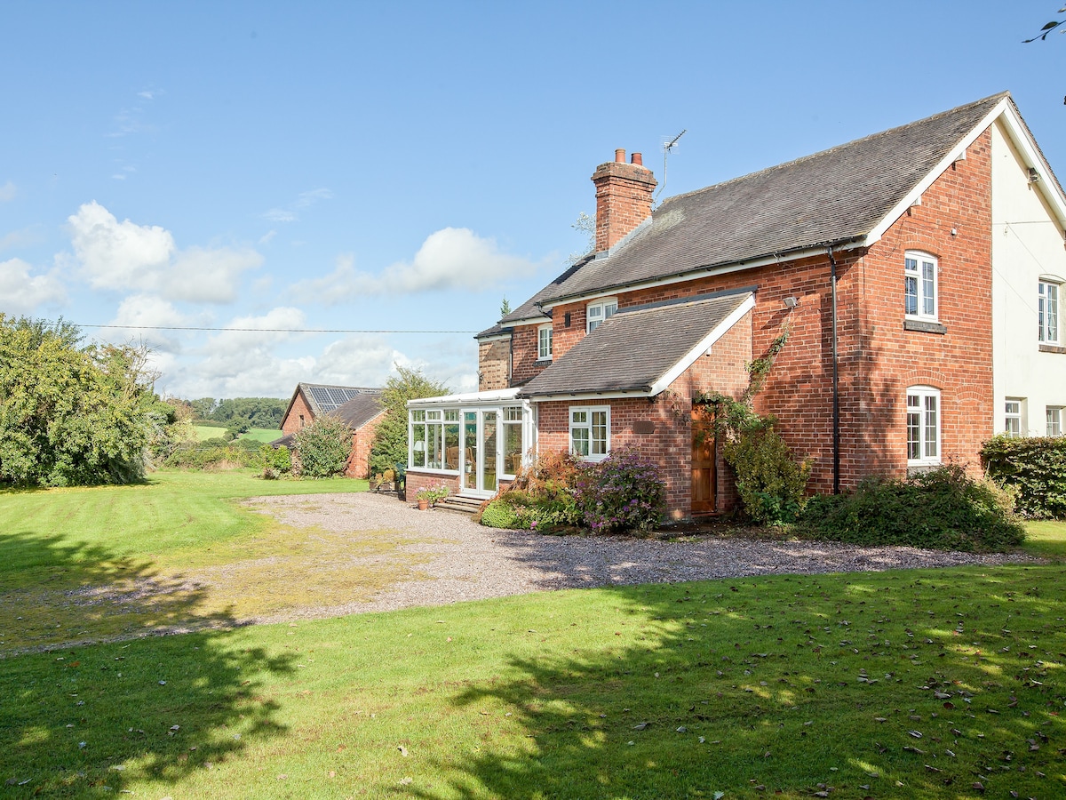A rural cottage is set in a spacious landscaped area, featuring a brick facade and a large conservatory. Lush greenery surrounds the property, with a gravel pathway leading to the entrance. The sky is clear with scattered clouds, highlighting the peaceful countryside setting.