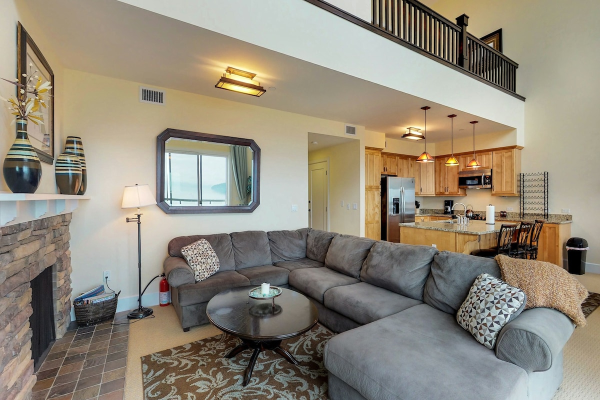A spacious living area is showcased, featuring a large gray sectional sofa arranged around a circular coffee table. A stone fireplace complements the space, while a full kitchen with wood cabinetry is visible in the background. Large windows provide natural light, enhancing the open atmosphere.