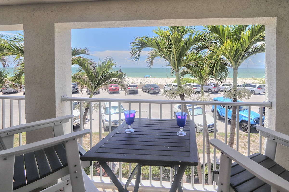 A private balcony is presented with outdoor seating, featuring a wooden table and two blue glasses. Lush palm trees frame the view, which highlights a sandy beach and the Gulf of Mexico beyond. Cars are visible parked along the street below.