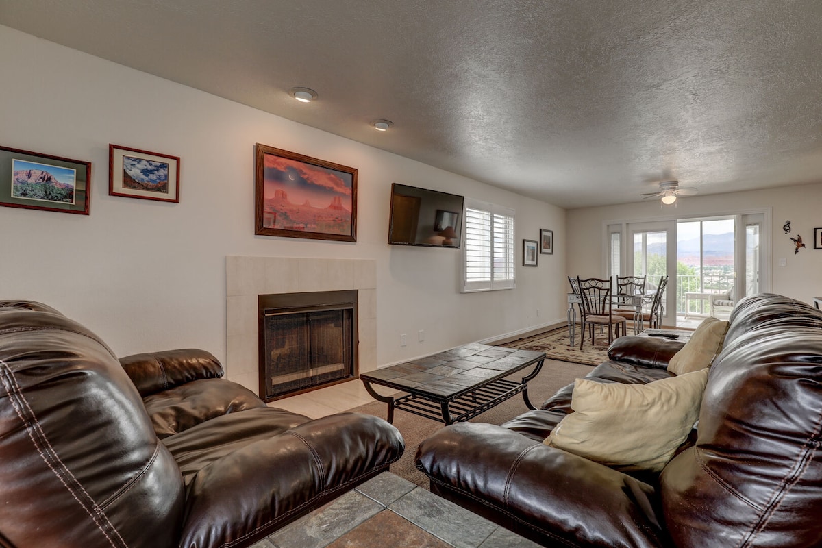 A spacious living area features comfortable leather seating arranged around a coffee table. A wall-mounted television is positioned above a fireplace. Natural light fills the room through sliding glass doors that lead to a balcony with mountain views, enhancing the overall inviting atmosphere.