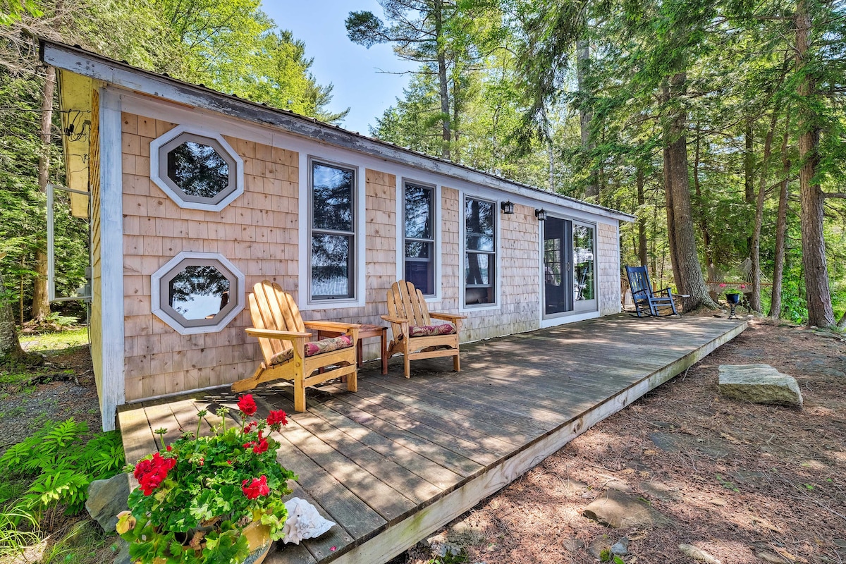 The cabin exterior features a spacious deck adorned with two wooden chairs, inviting views of the landscaped surroundings. Large windows and unique octagonal accents enhance the cabin's design, while colorful flowers and natural elements add charm to the serene setting.