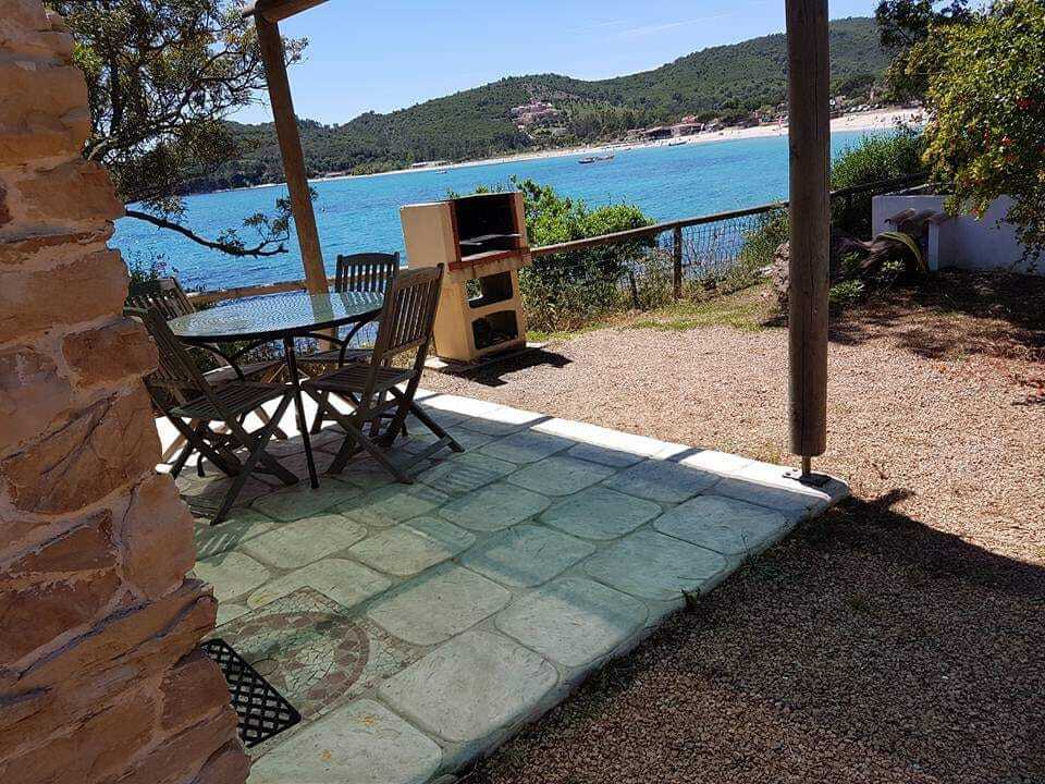 An outdoor terrace is visible, featuring a round table surrounded by folding chairs. A private barbecue is positioned nearby, with the bright blue sea and sandy beach in the background, framed by green foliage and hills.