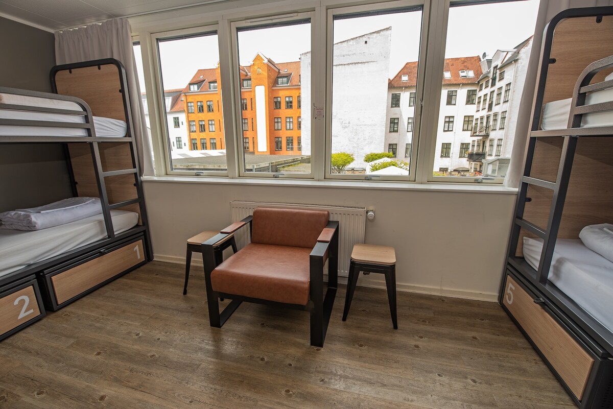 The room features two sets of bunk beds with individual bedding and under-bed lockers. A brown armchair is placed between two small black tables, facing large windows that offer views of city buildings and allow natural light to fill the space.