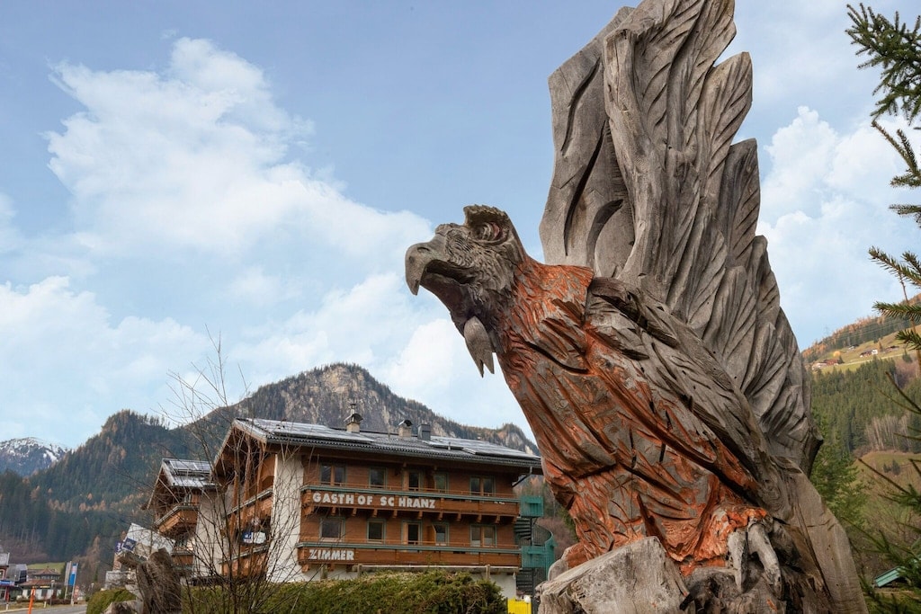 A prominently carved wooden sculpture of an eagle stands in front of a hotel building, showcasing intricate details in its feathers. Behind the sculpture, a mountainous landscape with soft clouds is visible, complementing the tranquil atmosphere of the surroundings.