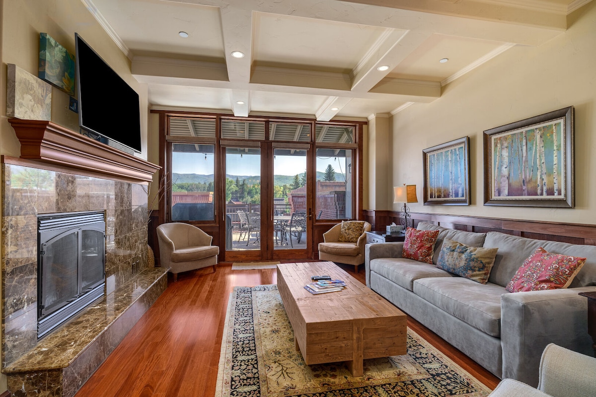 The living area features hardwood floors and a contemporary design, highlighted by a gas fireplace and a large flat-screen television. Large glass doors provide access to a spacious deck, while two comfortable armchairs and a sectional sofa with decorative cushions create a welcoming space.