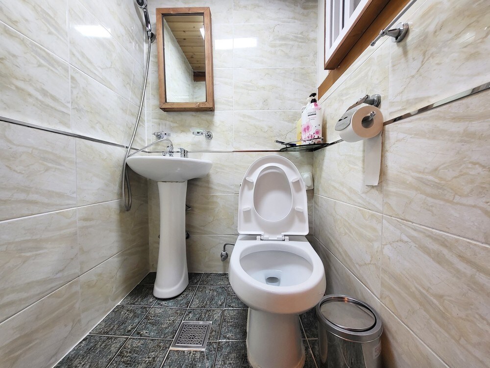 A clean and modern bathroom is presented, featuring a white toilet and a pedestal sink. Neutral-colored tiles cover the walls and floor. A small mirror is mounted above the sink, with toiletries neatly arranged on a shelf above. A waste bin is located beside the toilet.