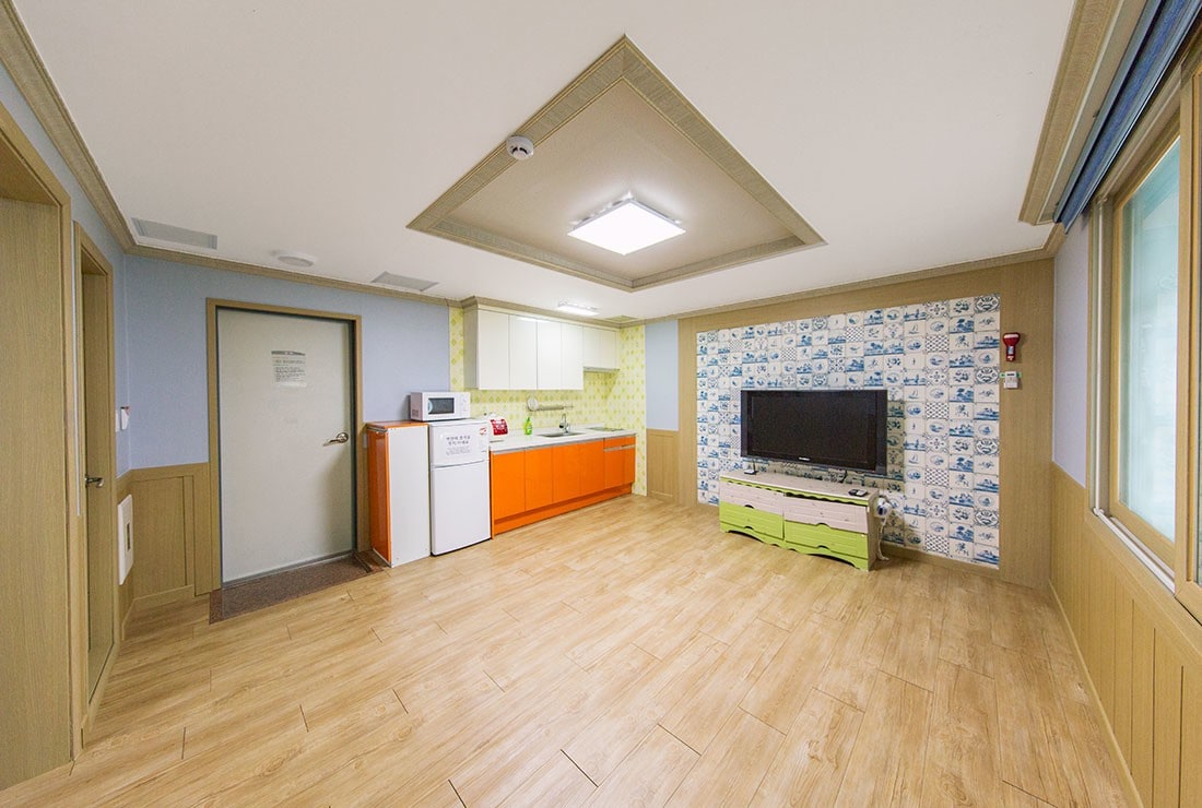 An open living space features an orange kitchen area with storage, a white washing machine, and a modern flat-screen television mounted on a wall. Large windows allow natural light to fill the room, enhancing the warm wooden flooring.