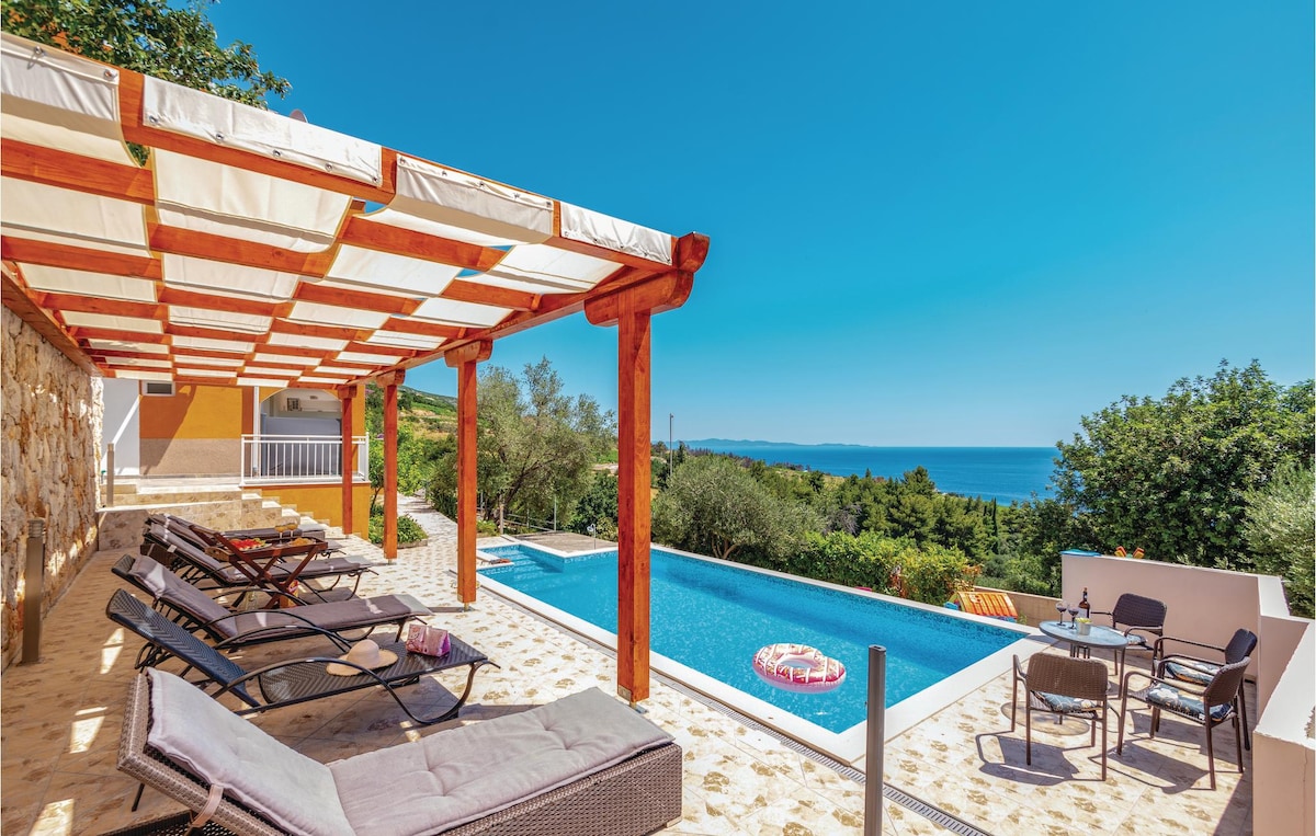 An outdoor terrace features a shaded area with reclining lounge chairs, overlooking a spacious pool. Lush greenery surrounds the pool area, providing a serene backdrop against the clear blue sky and distant sea view.