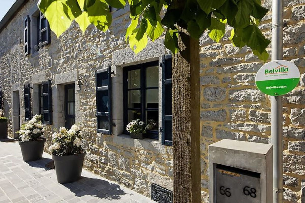 The exterior of the charming stone cottage is framed by vibrant green leaves and is flanked by large planters featuring blooming flowers. Windows with blue shutters add character, while a stone pathway leads to the entrance, hinting at a warm and inviting atmosphere.