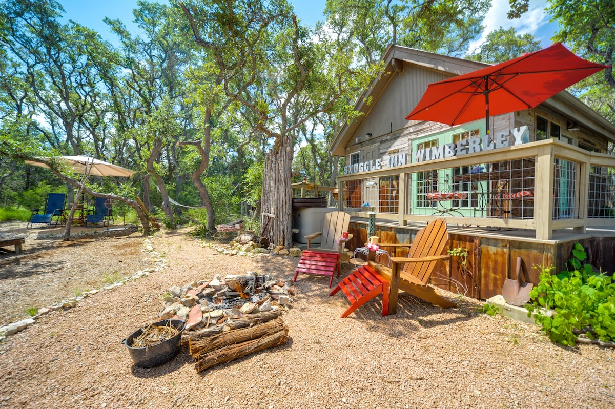 An outdoor area features a rustic cabin surrounded by live oak trees. A cozy fire pit is set with Adirondack chairs, while a shaded seating area with an umbrella provides a restful spot. A hammock is visible, inviting relaxation amid the natural surroundings.