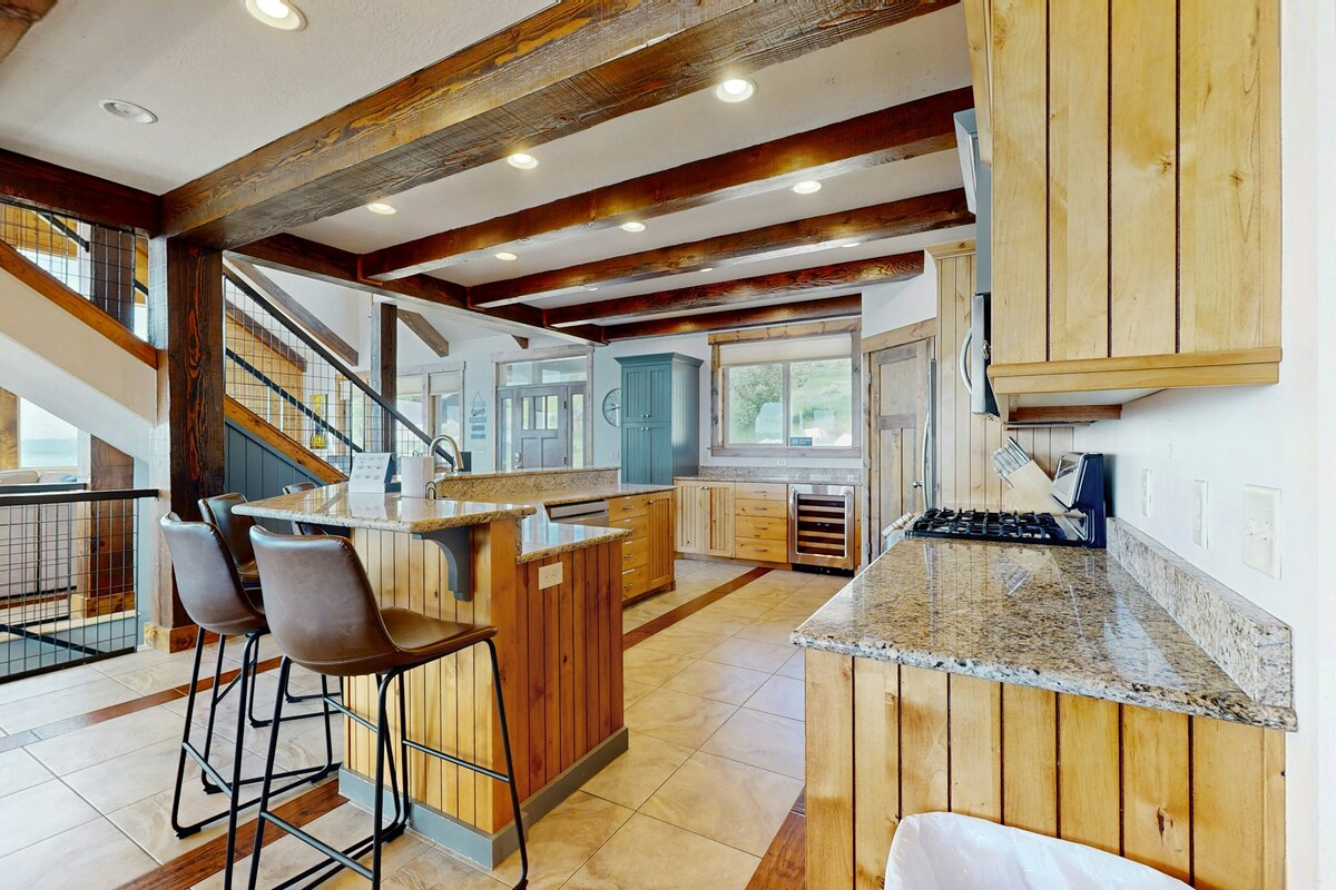 The kitchen features natural wood cabinetry and granite countertops, complemented by a spacious island with seating for three. Modern appliances are visible, and large windows provide natural light, illuminating the open layout with exposed wooden beams overhead.