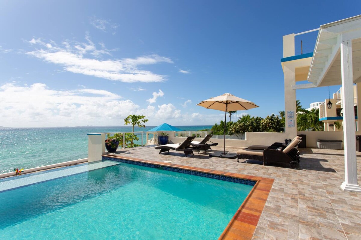An infinity pool features a clear blue surface that blends with the sea beyond. Poolside loungers and an umbrella provide shaded seating. The surrounding patio includes stone pavers, with a gentle view of the Caribbean waters and bright sky enhancing the serene setting.