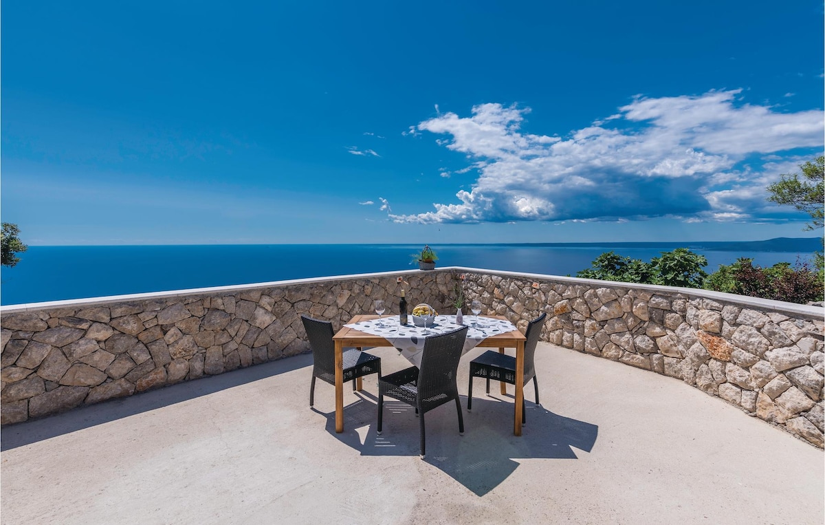 A spacious outdoor terrace features a round dining table surrounded by black chairs. The table is set with a vase and a plate, framed by a stone wall. A panoramic view of the blue sea and a clear sky enhances the serene atmosphere.