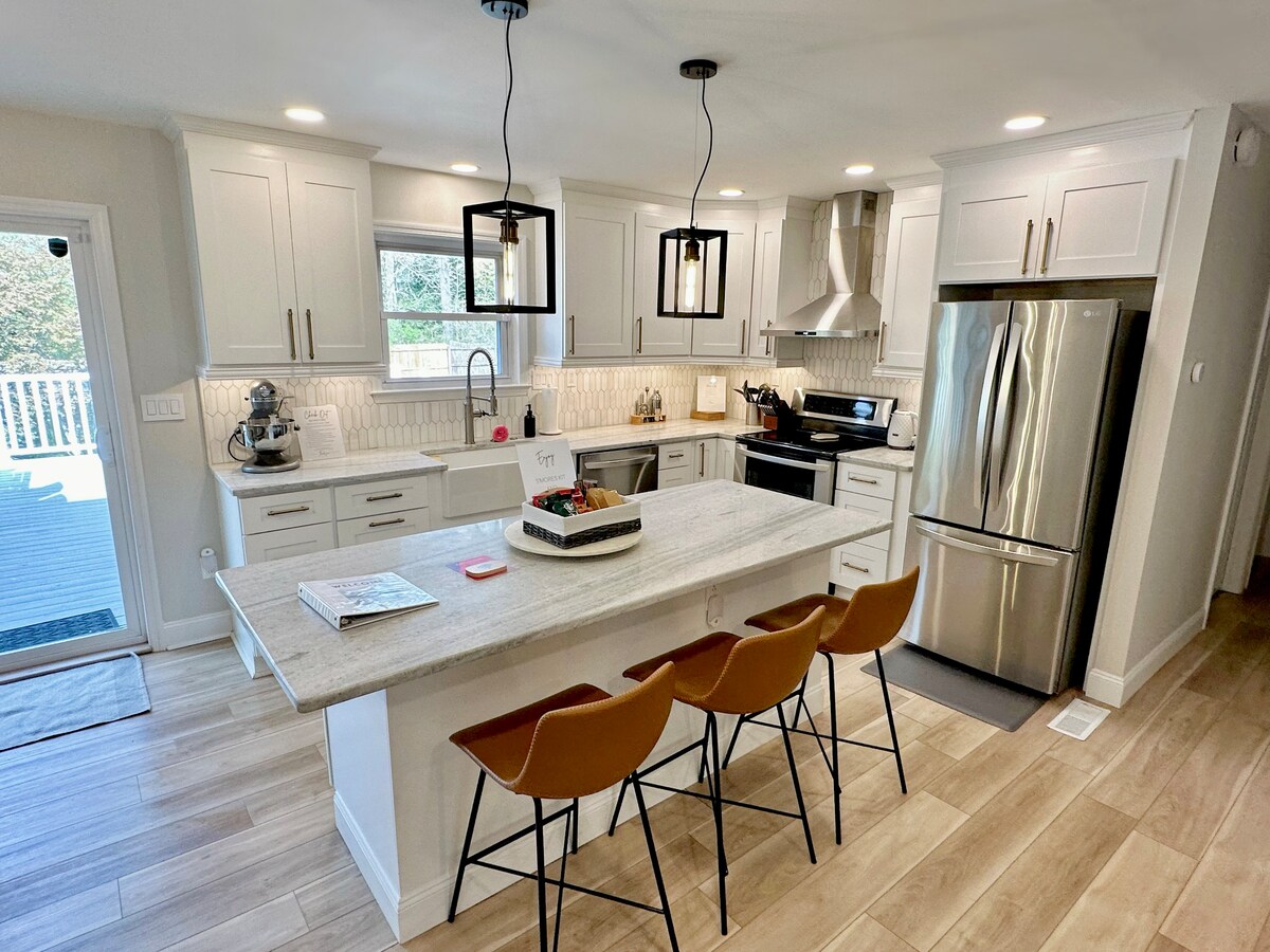 The kitchen features modern cabinetry and stainless steel appliances, including a refrigerator and oven. A spacious island with three bar stools provides additional seating. Natural light brightens the room through a nearby window, creating an inviting space for meal preparation.