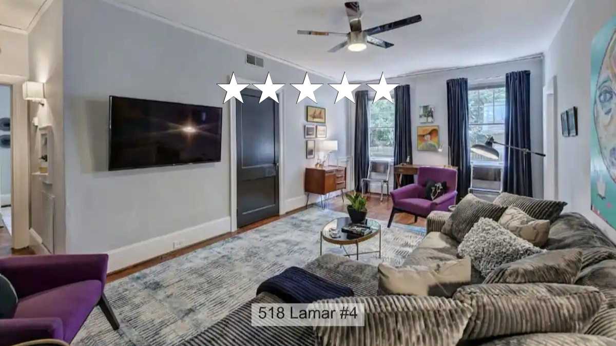 A spacious living room features a large, upholstered sofa accented with decorative pillows, alongside a plush armchair. A flatscreen television is mounted on one wall, while a modern ceiling fan offers airflow. Large windows dressed in blue curtains allow natural light to enhance the space.