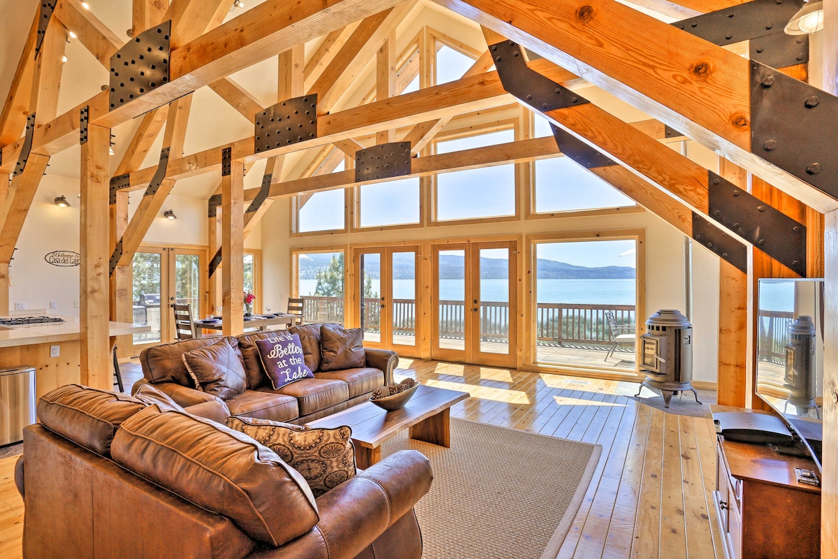 A spacious living area features vaulted ceilings with exposed wooden beams and large windows offering views of Eagle Lake. Comfortable seating is arranged around a coffee table, while a wood-burning stove adds a cozy touch. Natural light enhances the warmth of the wood accents.
