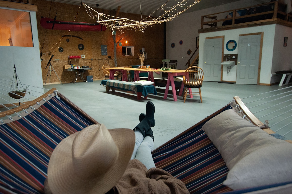 A spacious loft area is illuminated by soft string lights. A colorful hammock is positioned in the foreground, while a large harvest table with chairs is centrally located. The room features a rustic wood decor, with a kitchenette and various activity equipment visible in the background.