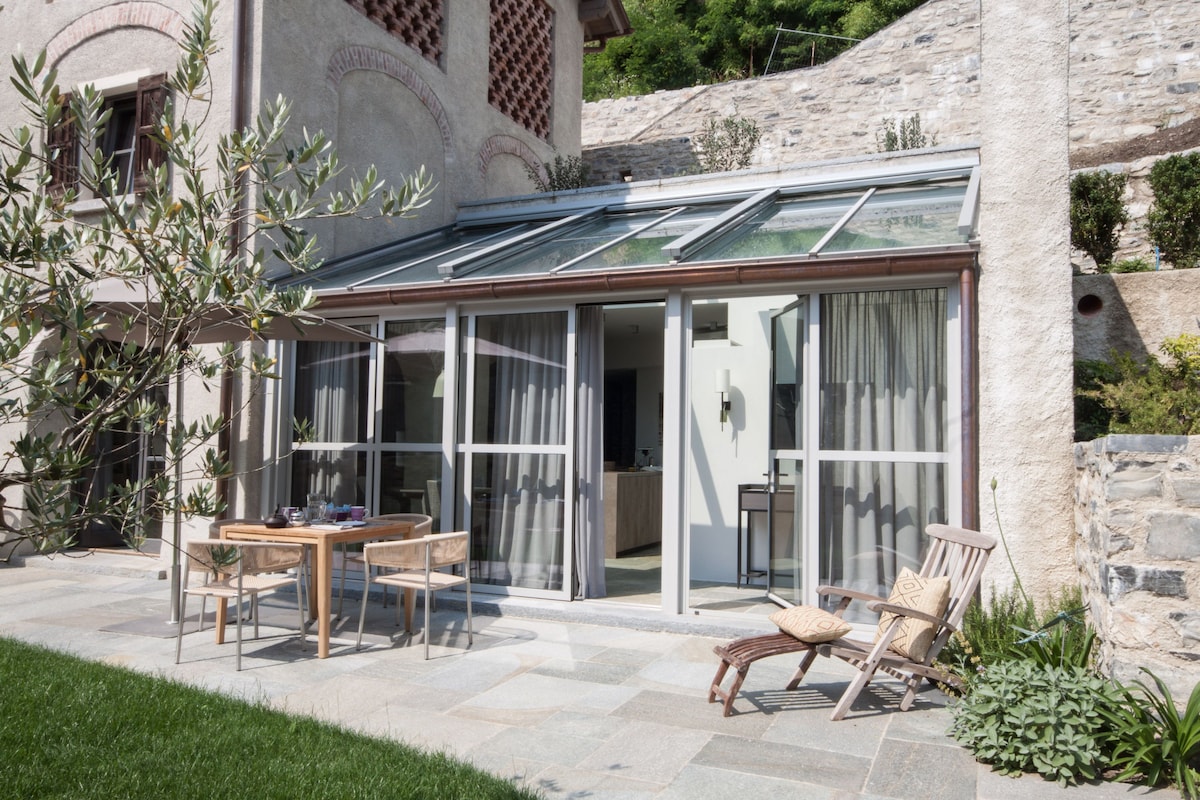 A sunlit terrace features a glass-roofed extension with large sliding doors. An outdoor dining table is set with chairs, while a pair of lounge chairs are positioned nearby. A well-maintained garden area and stone pathway complete the inviting outdoor space.