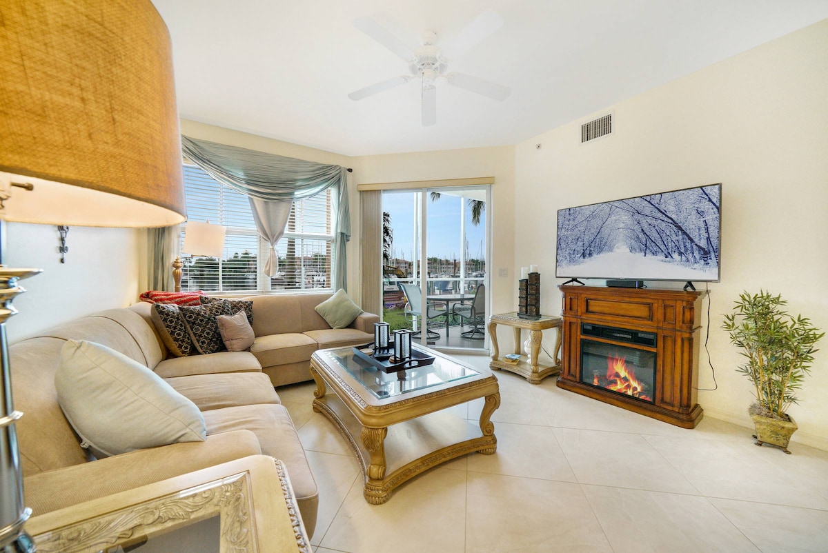 A comfortable living area is furnished with a spacious couch, a wooden coffee table, and a media console featuring a television. Natural light enters through large windows adorned with light drapes, and a decorative plant adds a touch of greenery. A cozy electric fireplace complements the space.