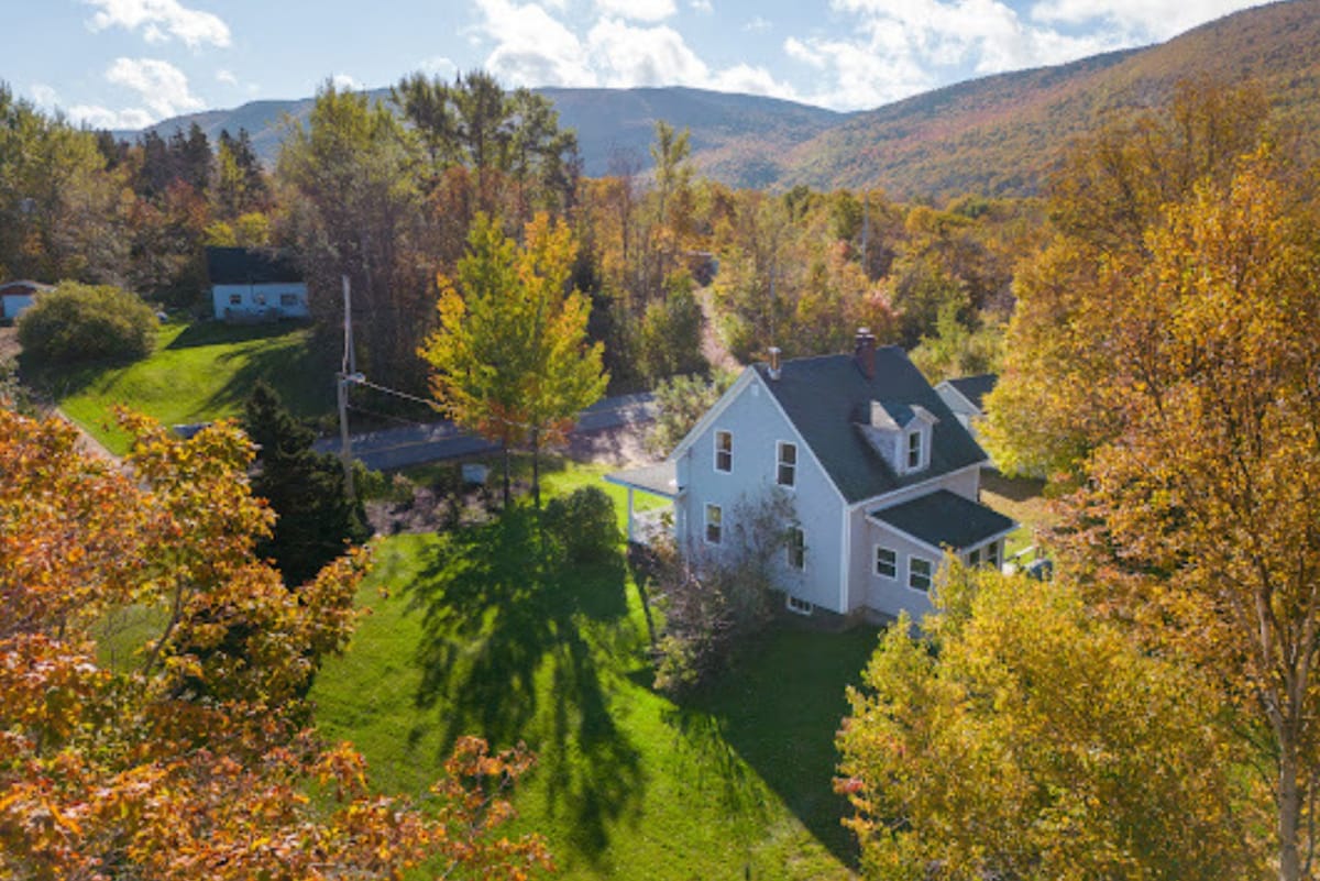 The cottage is surrounded by a spacious yard and vibrant trees showcasing autumn colors. The two-story structure features a sloped roof and is positioned near a winding road, with mountains visible in the background under a partly cloudy sky.