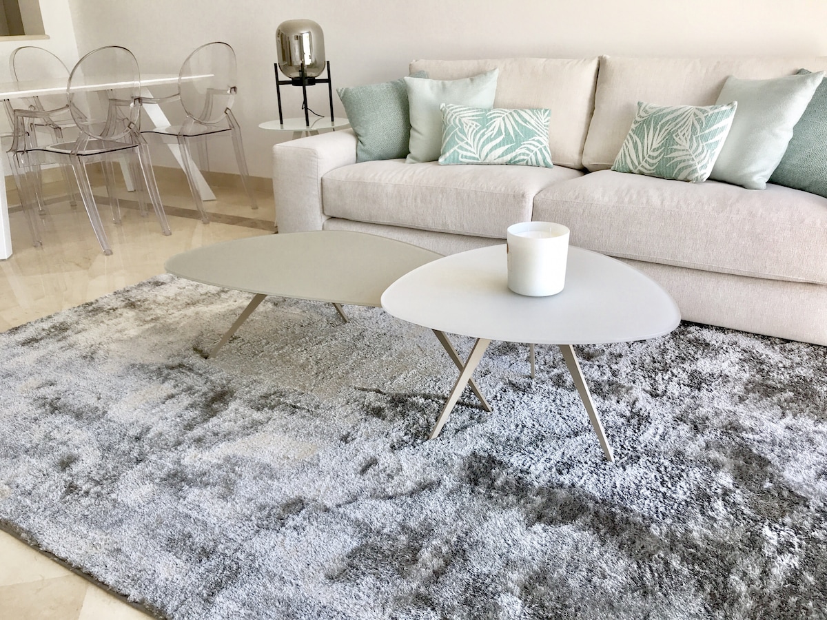A contemporary living area is present, featuring a light-colored sofa adorned with turquoise cushions. Two uniquely shaped coffee tables are positioned atop a textured grey rug, complementing the neutral tones. A dining area with transparent chairs is visible in the background.