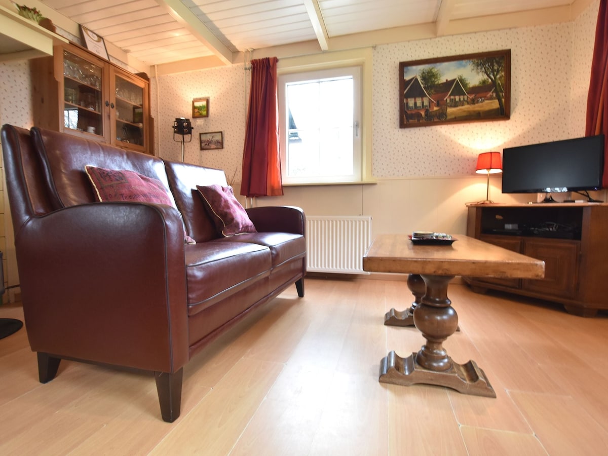 A living area is depicted, featuring a rich brown leather couch adorned with decorative pillows. A small wooden table is positioned in front, supported by a detailed pedestal base. An entertainment unit with a television is visible, complemented by warm lighting from a nearby lamp.