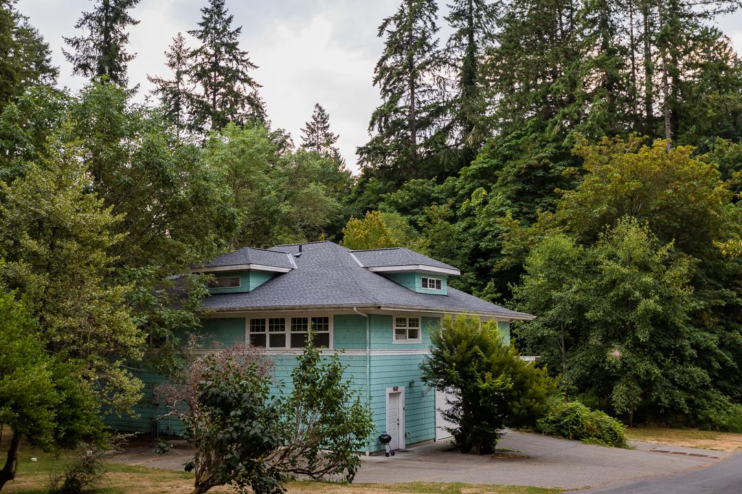 A two-story house is surrounded by lush greenery, featuring a light blue exterior. The sloped roof is adorned with darker shingles, and large windows provide views of the natural landscape. The driveway leads up to the entrance, nestled among trees.