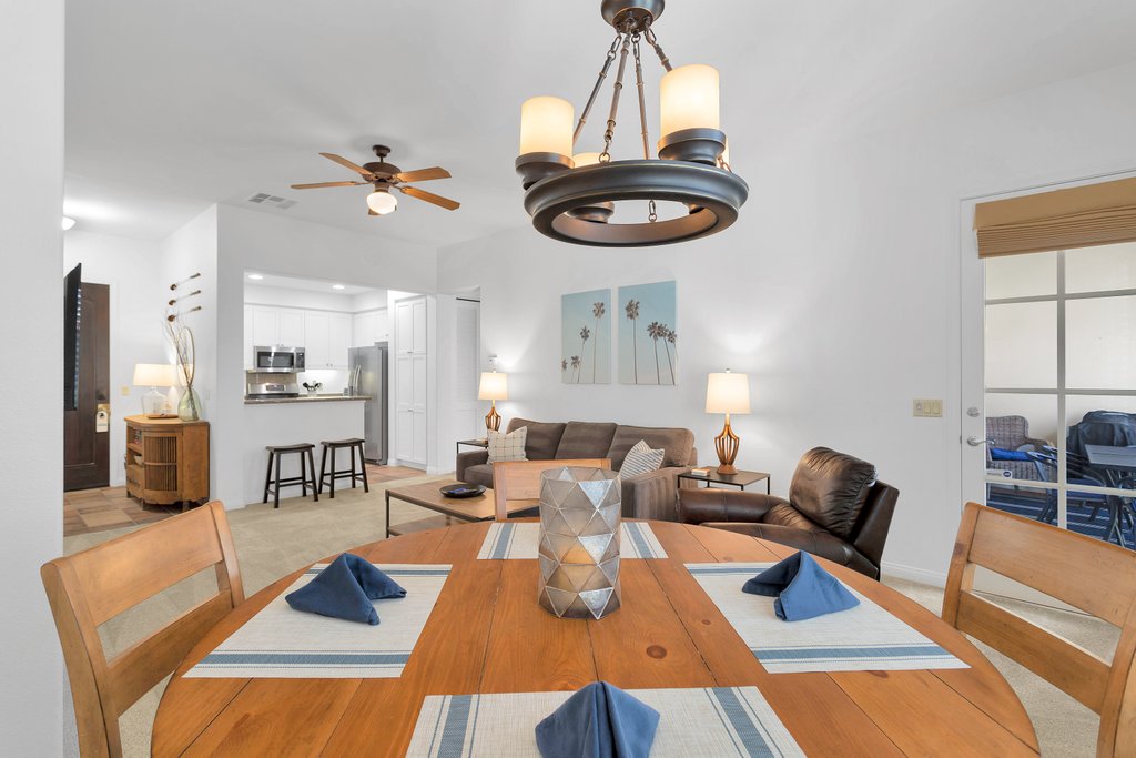 A dining area features a round wooden table set with blue napkins and a decorative centerpiece. A ceiling fan hangs above, and light from a chandelier highlights the space. In the background, a cozy living room with a sofa and chairs is visible, along with the kitchen area.