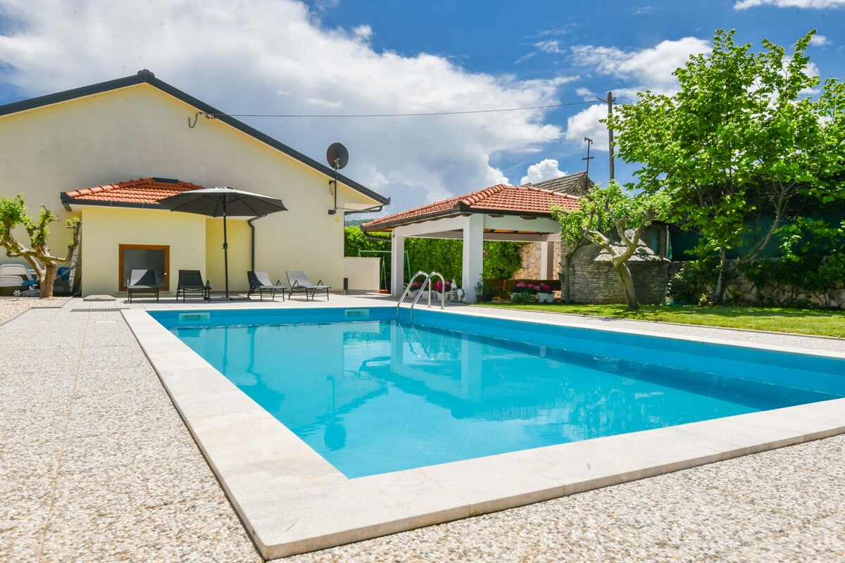 A private pool is set within a spacious terrace area, featuring sun loungers and a shaded seating arrangement. Lush greenery surrounds the pool, while a covered patio and home are visible in the background under a bright blue sky.