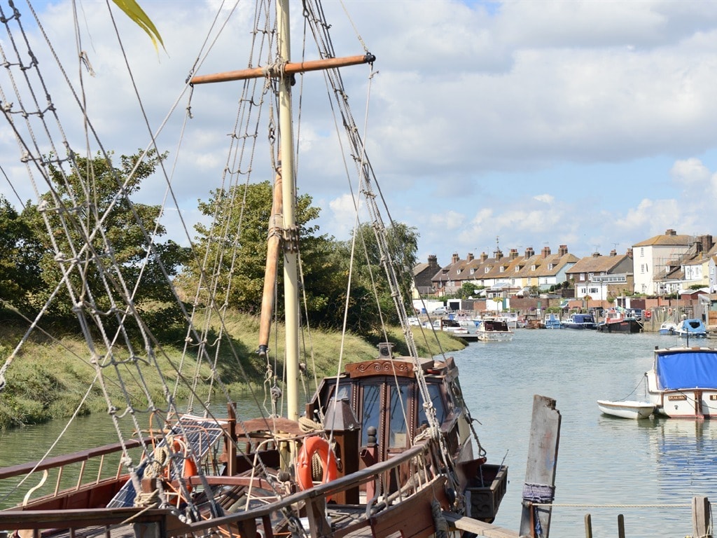 A wooden sailing ship is anchored along a tranquil waterway, with its tall mast and rigging prominently displayed. Lush green banks are visible on either side, leading to rows of quaint houses in the distance, all under a partly cloudy sky.